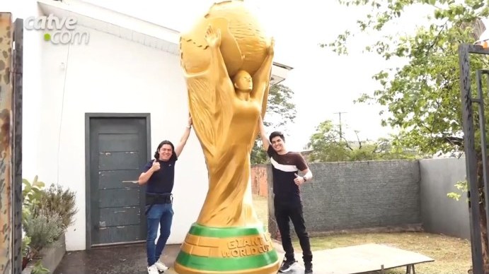 Giant golden World Cup replica in front of a house, two men posing next to it