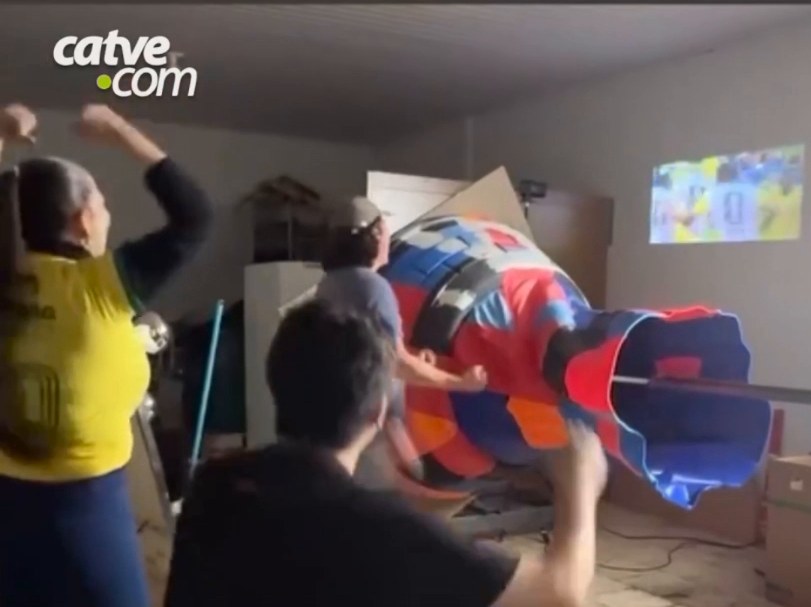 Multicoloured assembled trophy lying horizontally in the workshop while the team watches a match projected on the wall