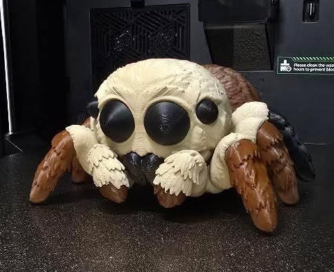 3D-printed jumping spider, beige head with large black eyes, brown and black legs, on a printer bed
